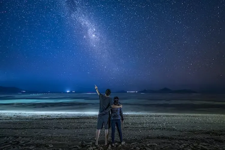 Two people standing on the beach during nighttime looking up at the stars.
